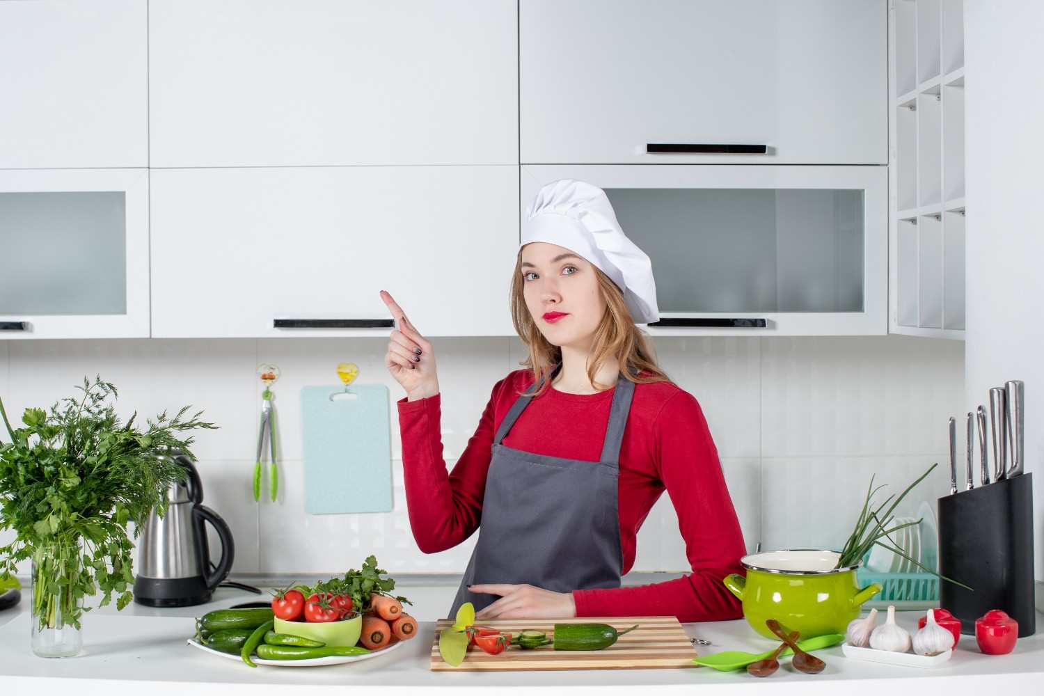 Female cook in a chef hat and apron pointing upward in a bright white kitchen