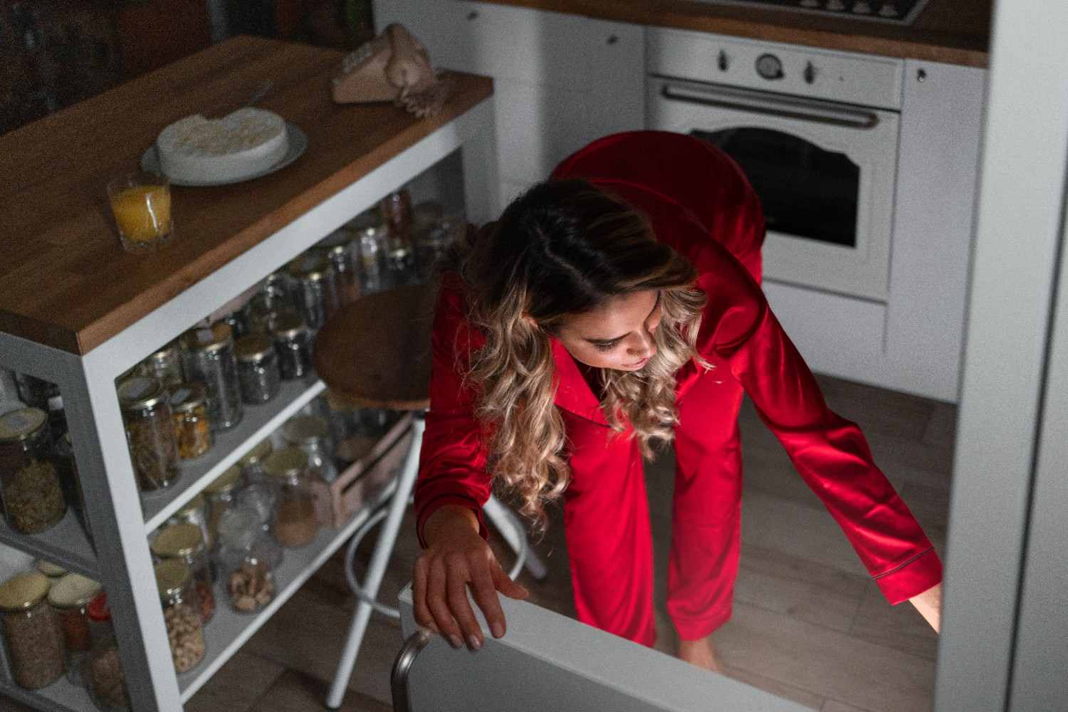 Woman in red pajamas looking into a fridge at night in a kitchen with jars of dry goods on open shelves